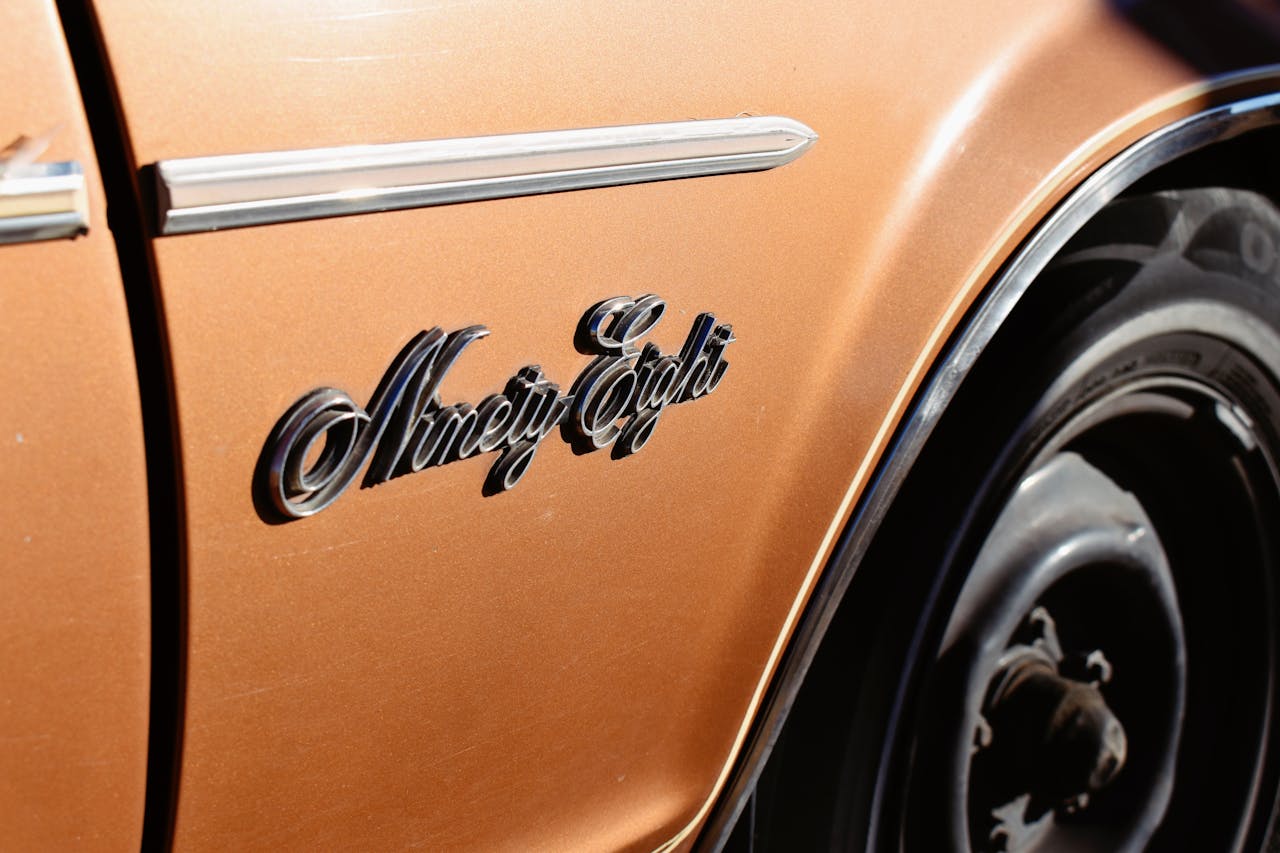 Primer plano de un emblema cromado de Oldsmobile Ninety-Eight en un coche clásico de color naranja.